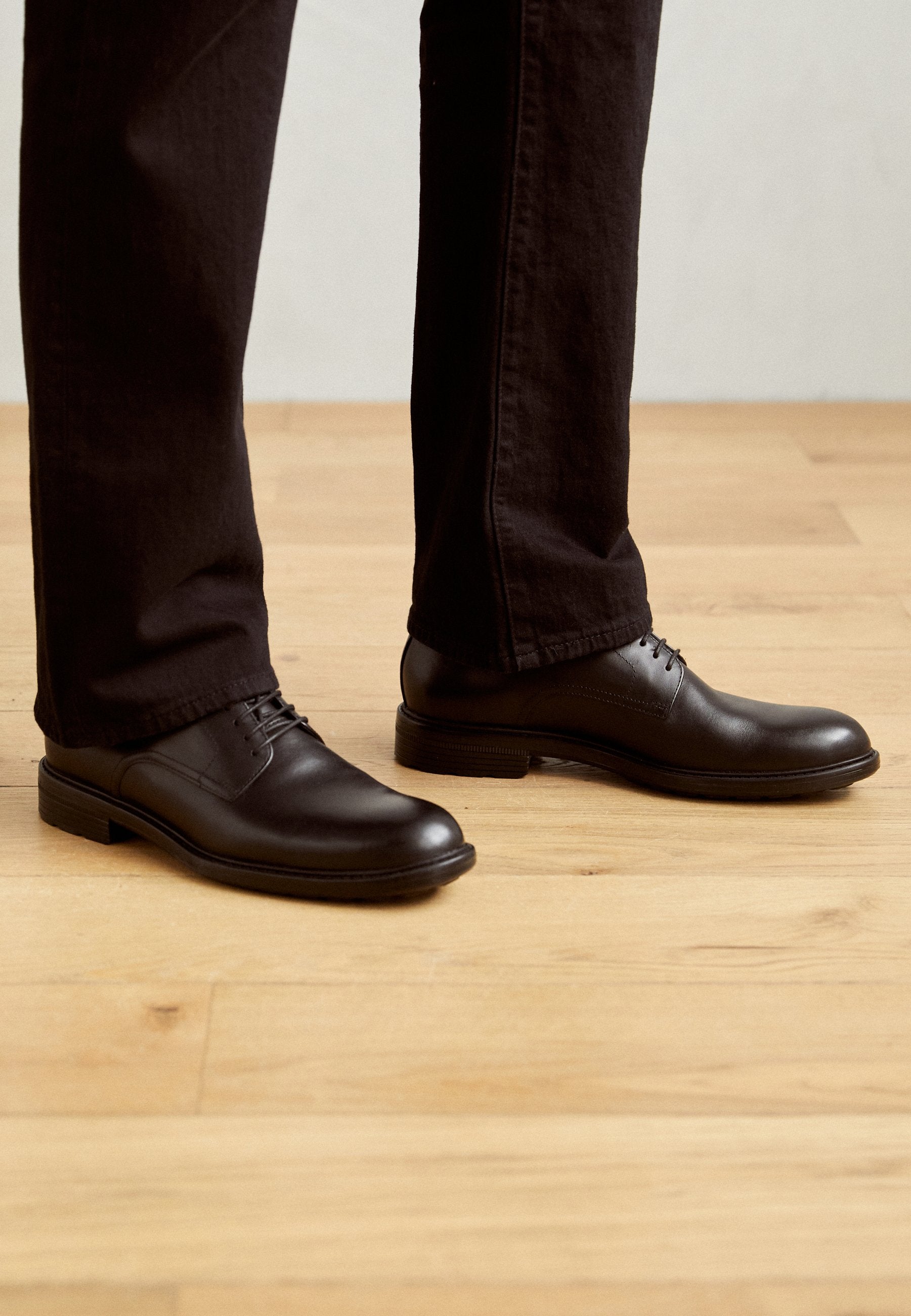 Schwarze Leder-Schnürschuhe mit glatter Oberfläche, abgerundetem Zeh und niedrigem Absatz, kombiniert mit dunkler Hose auf einem Holzboden.