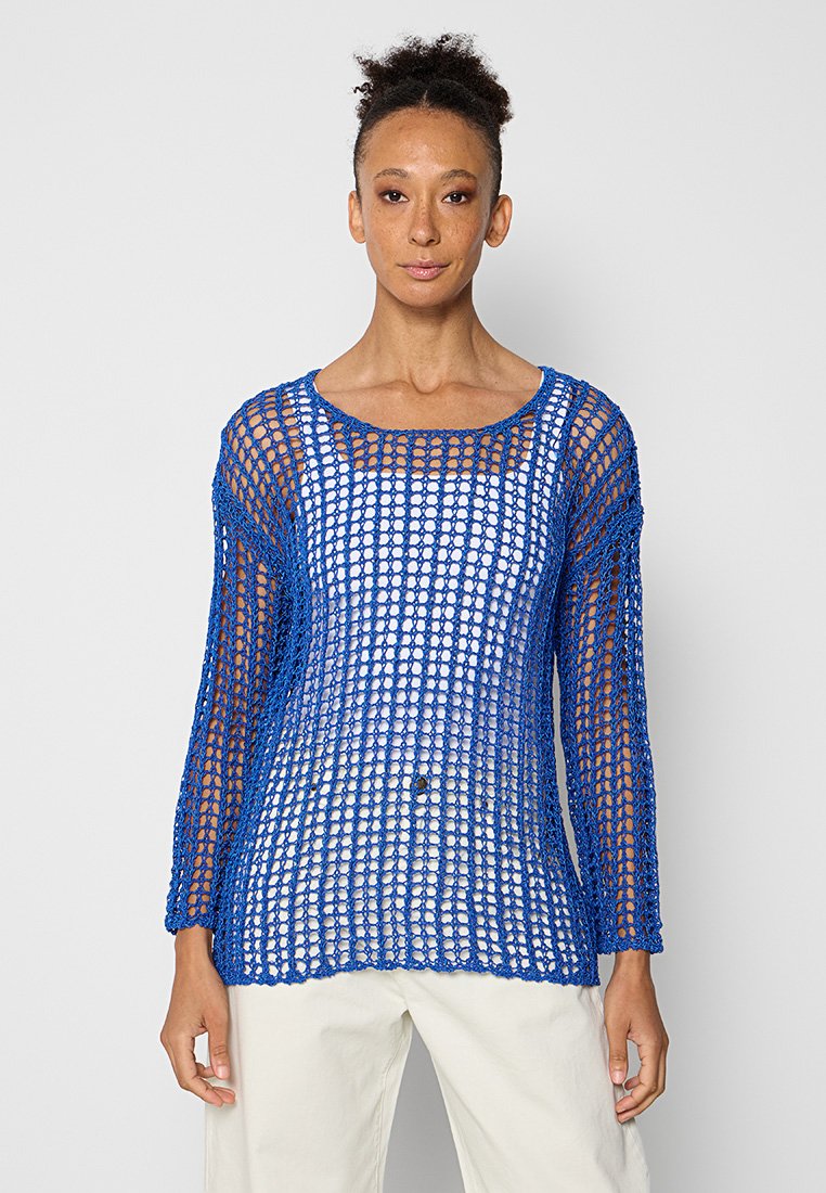 Eine Frau trägt einen blauen, offenen Strickpullover mit langen Ärmeln über einem weißen Oberteil und weißen Hosen. Sie steht vor einem schlichten, hellen Hintergrund.