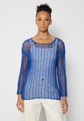 Eine Frau trägt einen blauen, offenen Strickpullover mit langen Ärmeln über einem weißen Oberteil und weißen Hosen. Sie steht vor einem schlichten, hellen Hintergrund.