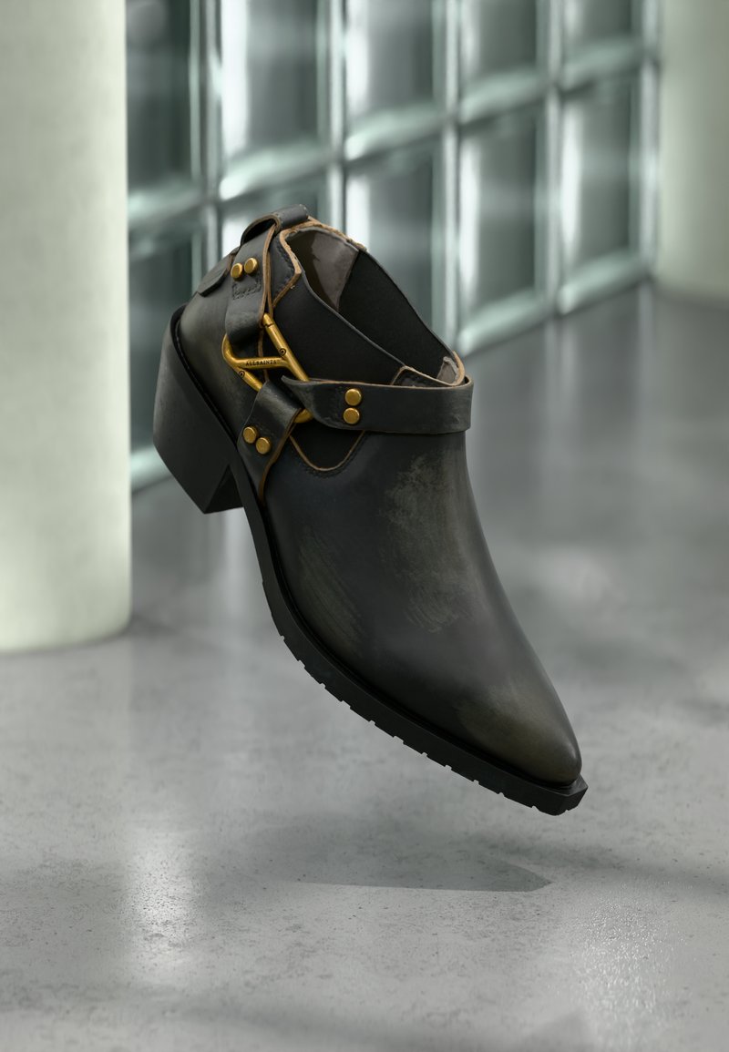 Schwarzer Lederschuh mit Absatz, goldener Schnalle und Riemendetails, platziert auf einem reflektierenden Betonboden vor einer Glasbausteinwand.
