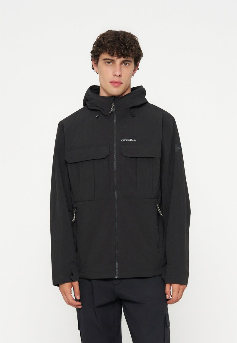 Junger Mann trägt eine schwarze Kapuzenjacke mit Frontreißverschluss, Klapptaschen und O'Neill-Logo auf der Brust, steht vor weißem Hintergrund.