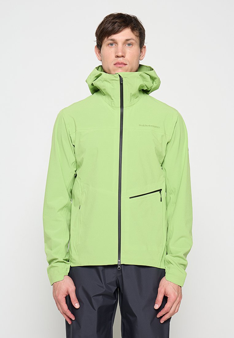 Mann steht und trägt eine hellgrüne Kapuzenjacke mit Frontreißverschluss und schwarzer Reißverschlusstasche, kombiniert mit dunkler Hose, neutraler Hintergrund.
