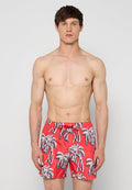 Oberkörperfreier Mann mit kurzen dunklen Haaren, der rote Badeshorts mit schwarzen und weißen Palmenmustern trägt und vor einem schlichten hellen Hintergrund steht.