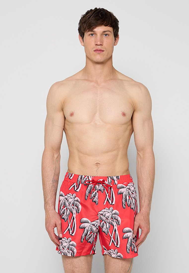 Oberkörperfreier Mann mit kurzen dunklen Haaren, der rote Badeshorts mit schwarzen und weißen Palmenmustern trägt und vor einem schlichten hellen Hintergrund steht.