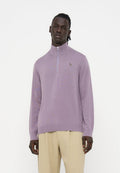Mann trägt einen hellvioletten Pullover mit Viertelreißverschluss und beige Hose, steht vor einem einfachen weißen Hintergrund.