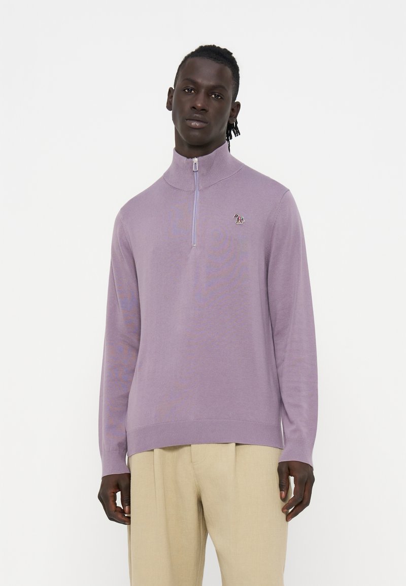 Mann trägt einen hellvioletten Pullover mit Viertelreißverschluss und beige Hose, steht vor einem einfachen weißen Hintergrund.