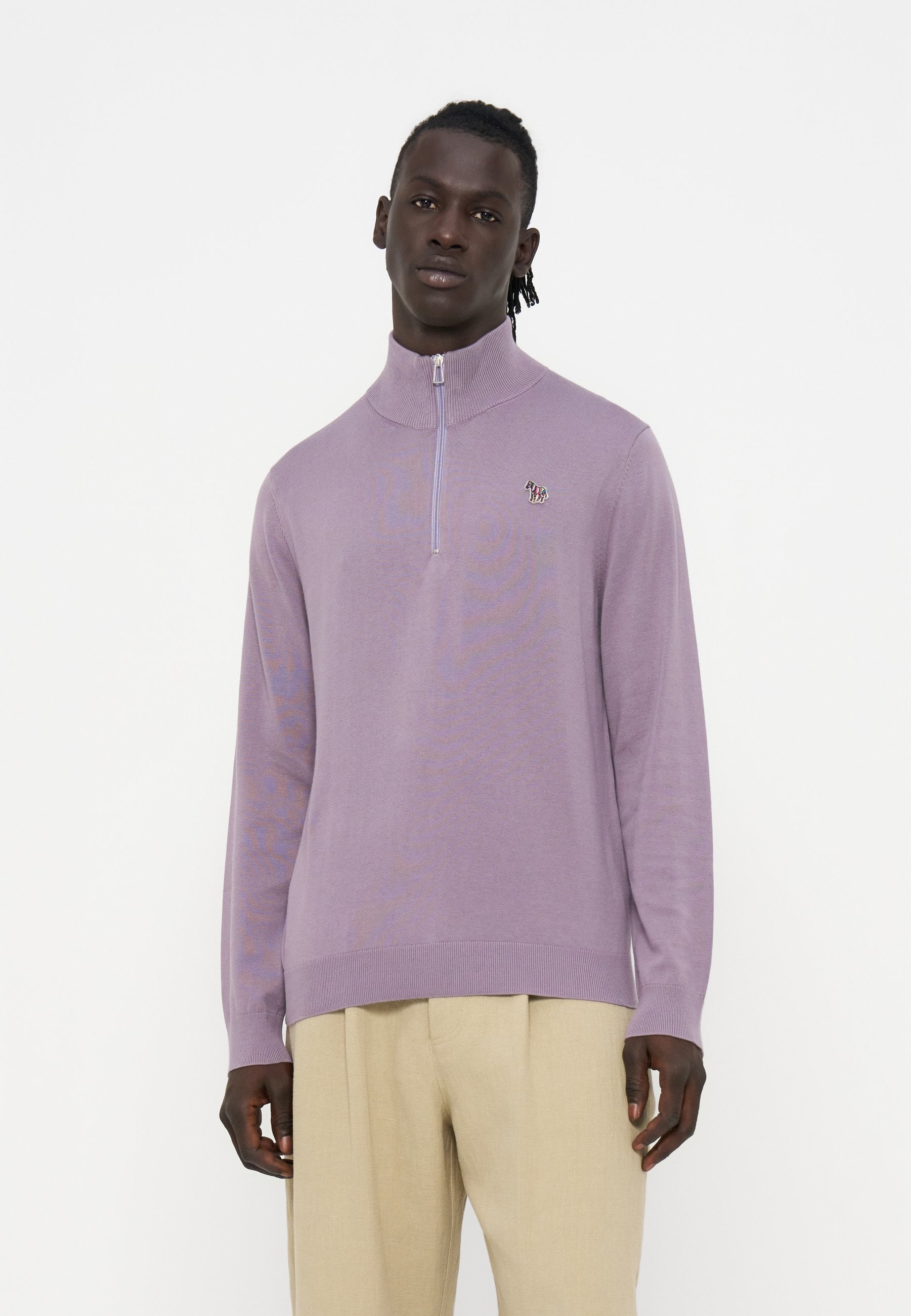 Mann trägt einen hellvioletten Pullover mit Viertelreißverschluss und beige Hose, steht vor einem einfachen weißen Hintergrund.