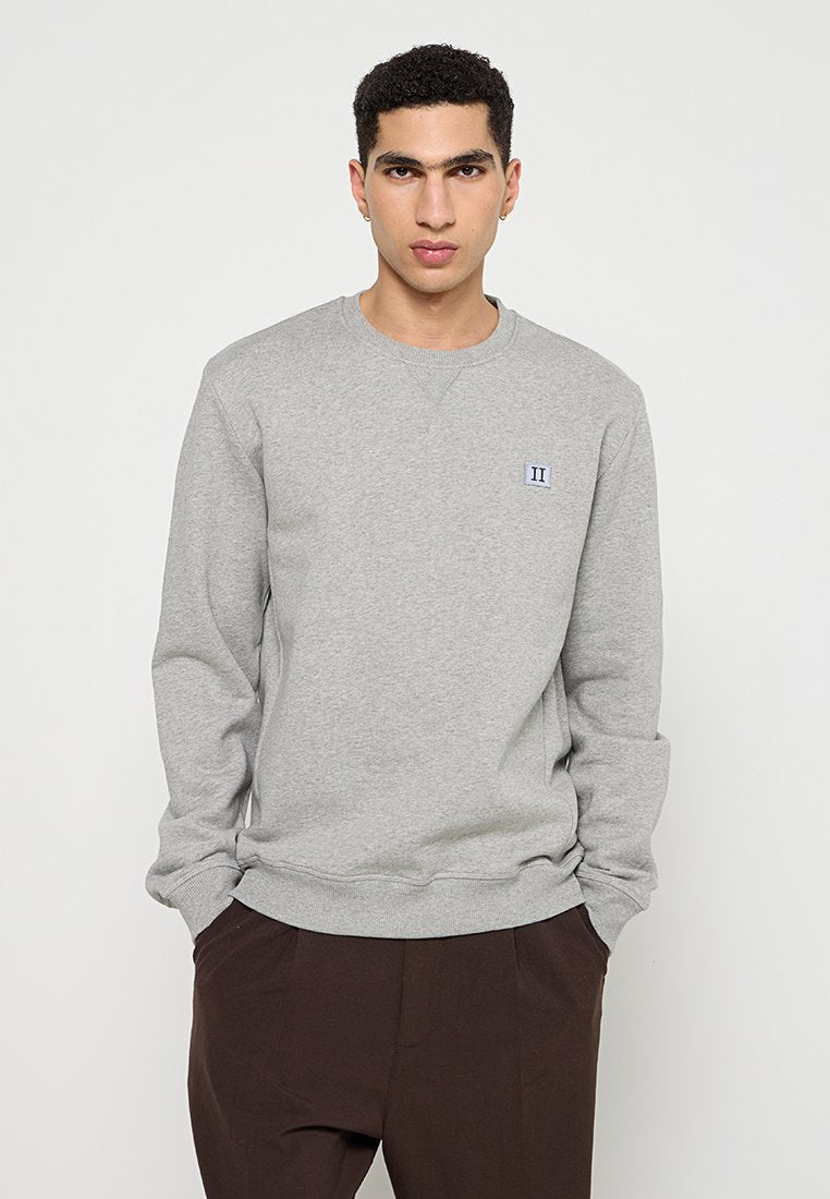 Junger Mann mit kurzen lockigen Haaren, der einen hellgrauen Sweatshirt mit einem kleinen Aufnäher auf der Brust und einer dunkelbraunen Hose trägt, mit den Händen in den Taschen.