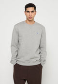 Junger Mann mit kurzen lockigen Haaren, der einen hellgrauen Sweatshirt mit einem kleinen Aufnäher auf der Brust und einer dunkelbraunen Hose trägt, mit den Händen in den Taschen.