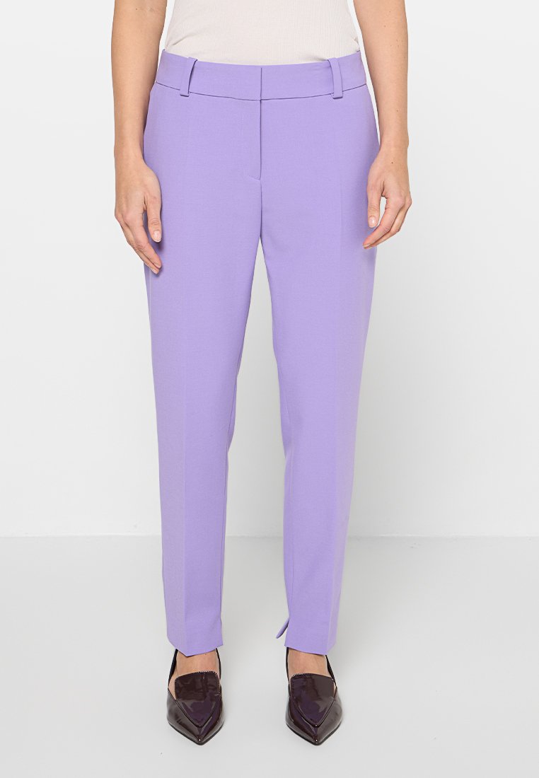 Fliederfarbene, maßgeschneiderte Hose aus glattem Stoff mit schmaler Passform, Gürtelschlaufen und seitlichen Taschen. Kombiniert mit dunklen spitzen Schuhen.