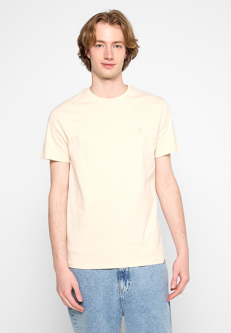 Hellbeige T-Shirt mit kurzen Ärmeln aus Baumwolle. Einfacher runder Ausschnitt, lockere Passform, mit einem kleinen erhabenen Logo auf der Brust.