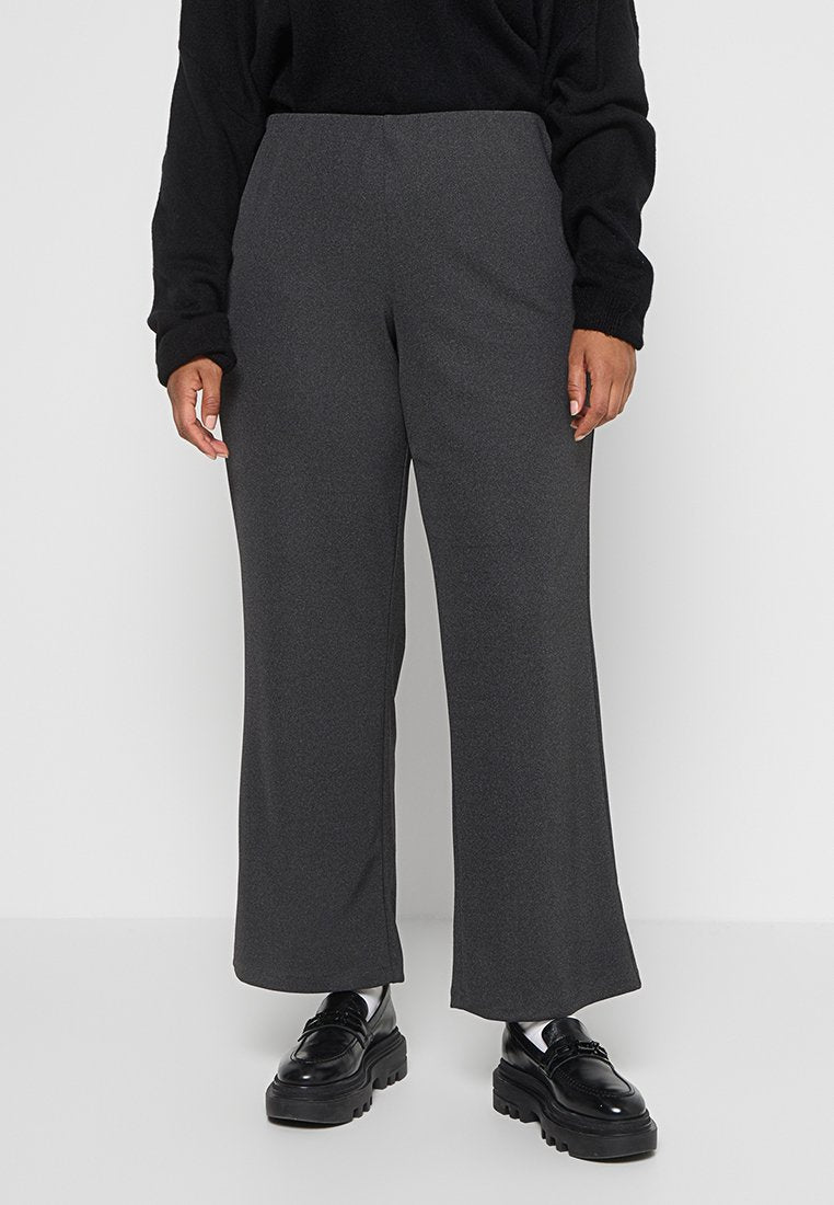 Person trägt dunkelgraue weit geschnittene Hose, schwarzes Langarmoberteil, weiße Socken und schwarze Plattformslipper und steht auf einem weißen Boden.