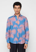 Mann mit kurzem lockigem Haar, der ein blau-rosa Batik-Langarmhemd mit Knöpfen und eine schwarze Hose trägt, steht vor einem schlichten Hintergrund.