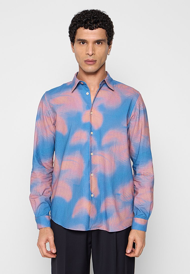 Mann mit kurzem lockigem Haar, der ein blau-rosa Batik-Langarmhemd mit Knöpfen und eine schwarze Hose trägt, steht vor einem schlichten Hintergrund.