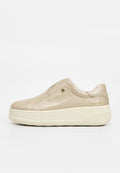 Beiger Slip-On-Ledersneaker mit dicker weißer Sohle und kleiner runder Metallverzierung nahe dem Knöchel auf einfarbigem Hintergrund.