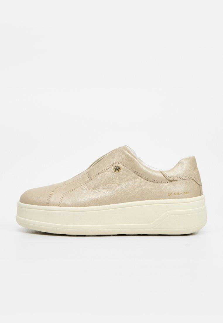 Beiger Slip-On-Ledersneaker mit dicker weißer Sohle und kleiner runder Metallverzierung nahe dem Knöchel auf einfarbigem Hintergrund.