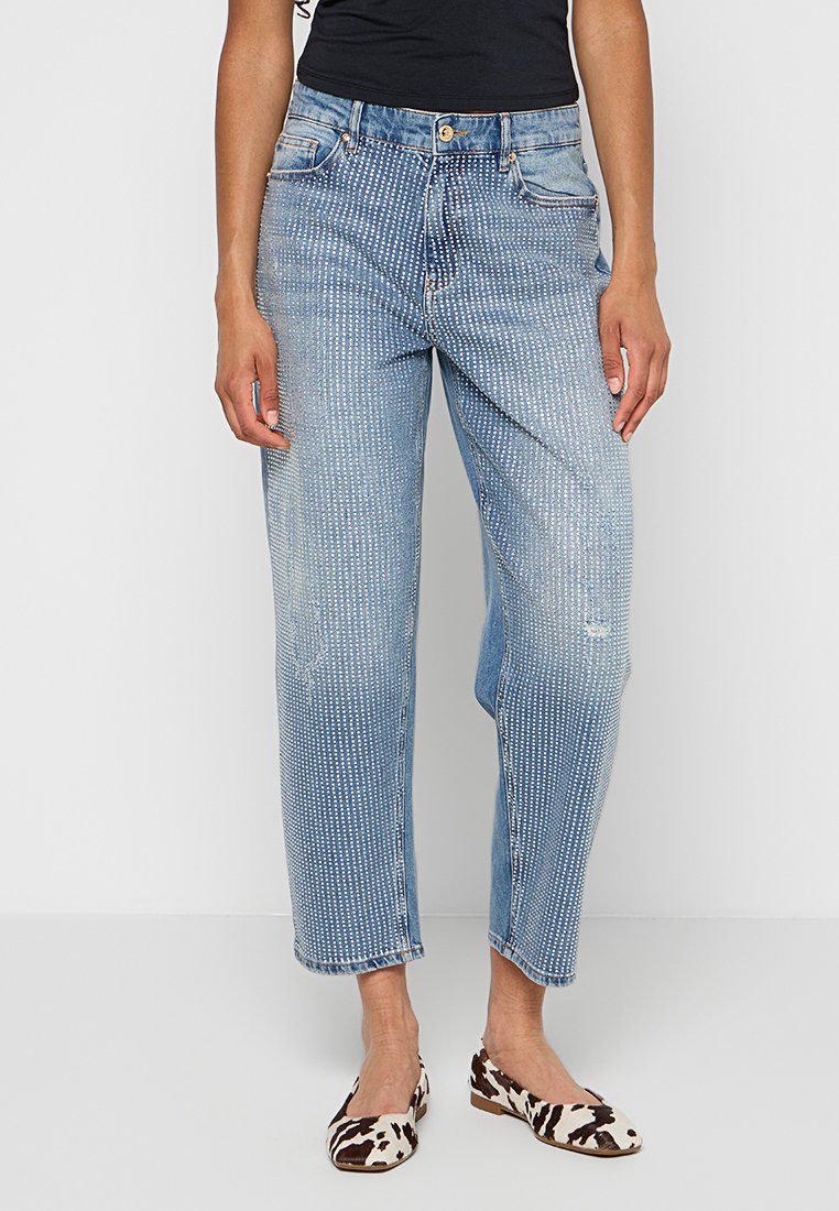 Hellblaue Denim-Jeans mit entspannter Passform, mit weißen gestickten Mustern, fünf Taschen und einem Knopfverschluss. Getragen mit gemusterten Ballerinas.