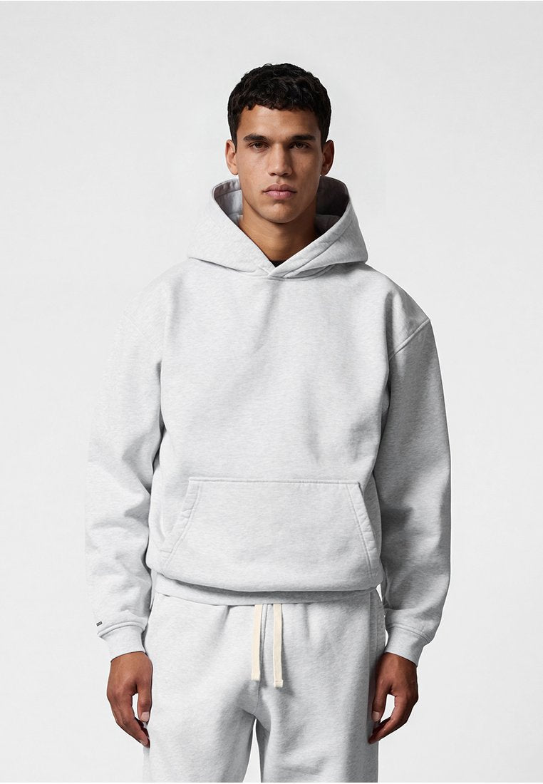 Junger Mann trägt hellgrauen Hoodie und passende Jogginghose, steht vor einem schlichten hellen Hintergrund, blickt frontal mit neutralem Gesichtsausdruck.
