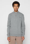 Junger Mann, der einen grauen Sweatshirt mit Viertelreißverschluss und kleinem Logo auf der Brust sowie beige Hosen trägt, steht vor einem schlichten Hintergrund.