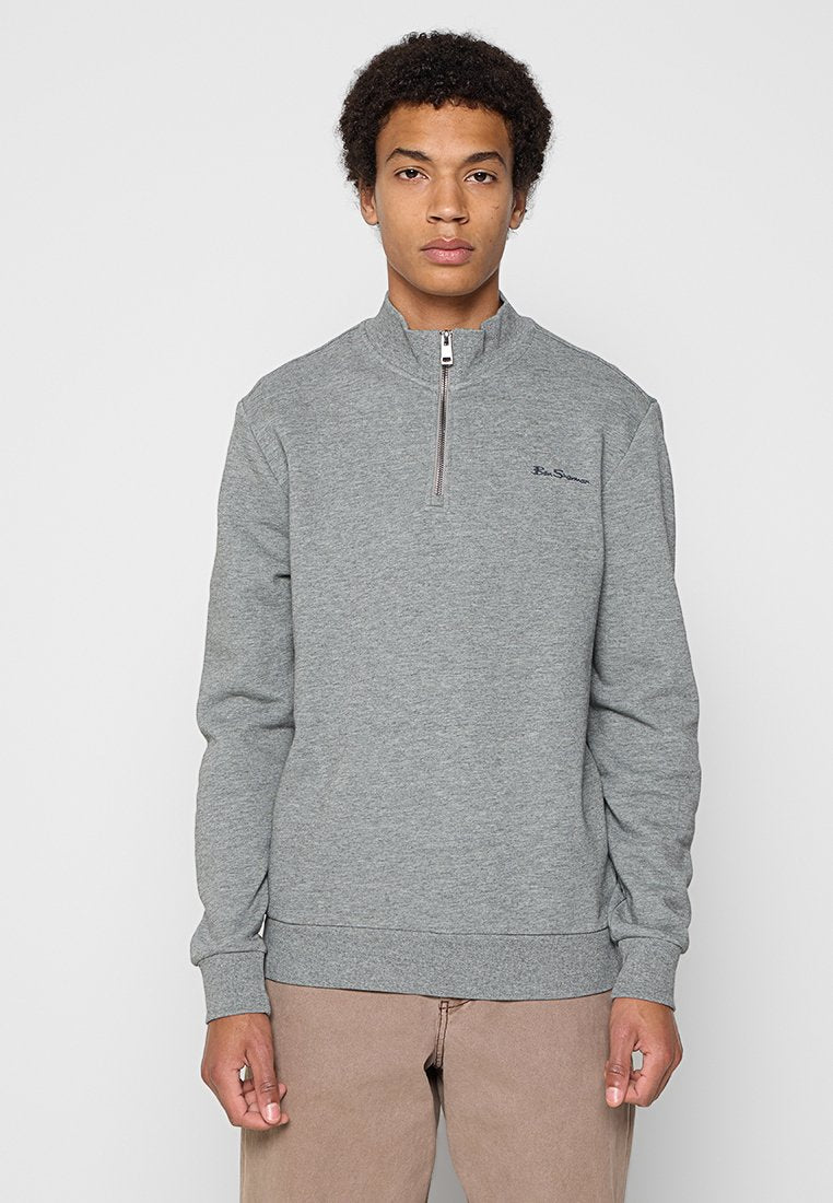 Junger Mann, der einen grauen Sweatshirt mit Viertelreißverschluss und kleinem Logo auf der Brust sowie beige Hosen trägt, steht vor einem schlichten Hintergrund.