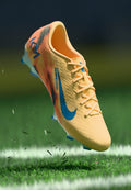 Helle gelbe Fußballschuhe mit orangefarbenen und blauen Akzenten, strukturiertem Obermaterial und blauem Nike-Swoosh; ausgestattet mit geformten Stollen für besseren Halt.