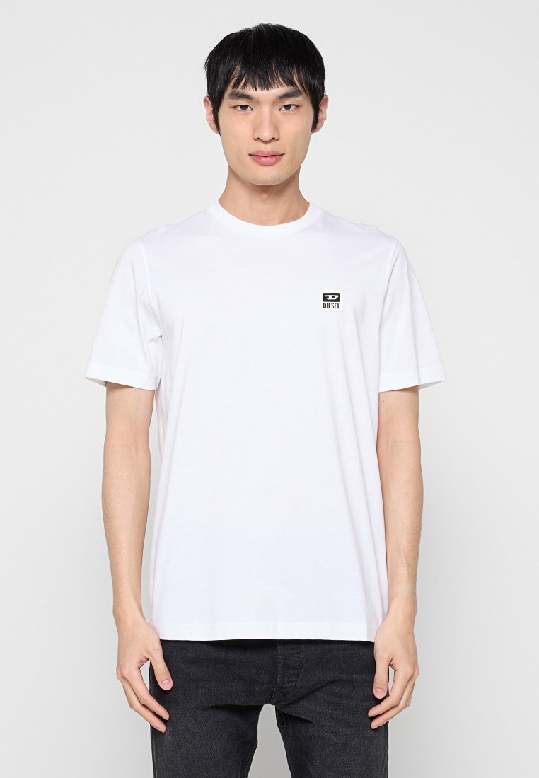 Weißes Baumwoll-T-Shirt mit kurzen Ärmeln, Rundhalsausschnitt und einem kleinen schwarzen Diesel-Logo-Patch auf der linken Brust. Kombiniert mit dunklen Jeans.