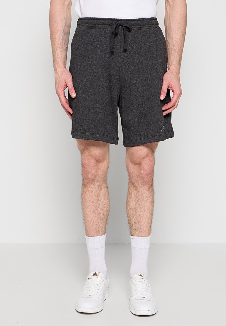 Mann trägt dunkelgraue Shorts mit Kordelzug, weiße Sportsocken und weiße Sneakers, steht vor neutralem Hintergrund.