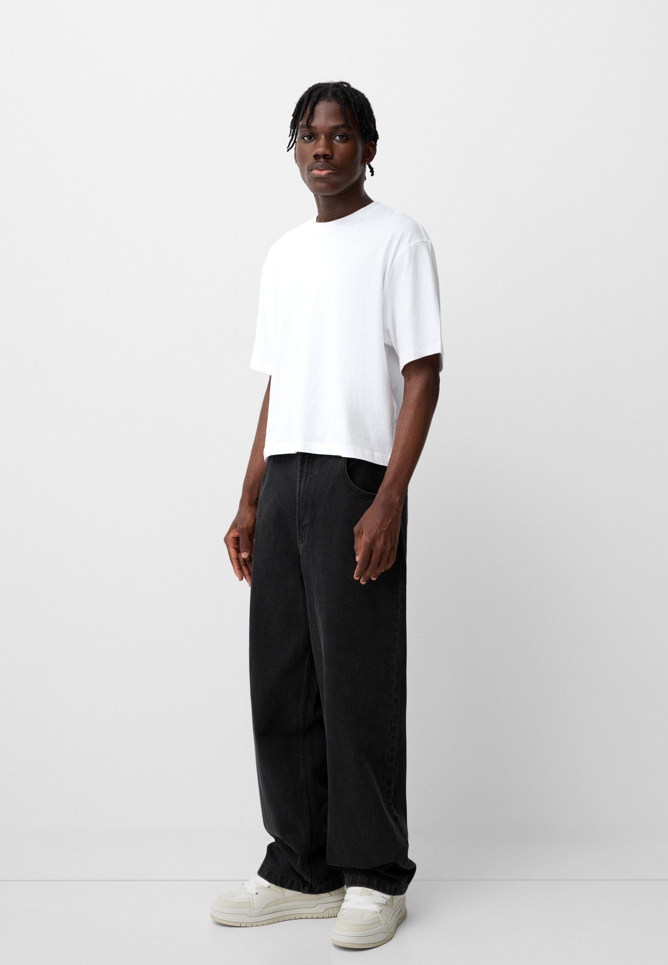 Weißen Baumwoll-T-Shirt, lockere Passform, kurze Ärmel. Schwarze Jeans mit weitem Bein, vordere Taschen. Helle Sneakers.