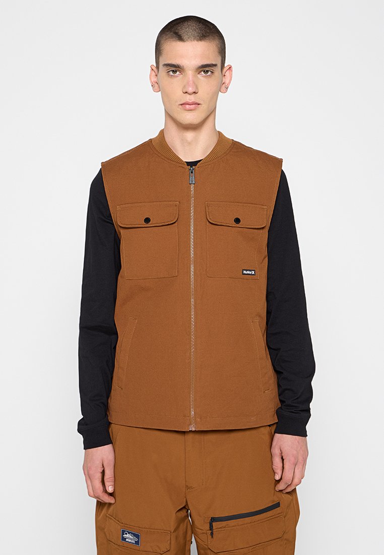 Junger Mann mit kurzen Haaren, der ein braunes ärmelloses Oberteil über einem schwarzen Langarmshirt und passenden braunen Cargo-Hosen mit Taschen trägt.