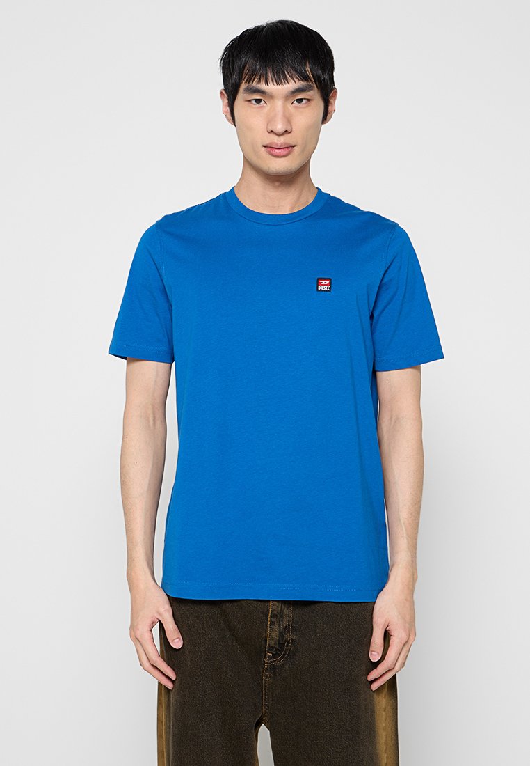 Blaues Baumwoll-T-Shirt mit rundem Halsausschnitt, kurzen Ärmeln und einem kleinen roten Logo-Patch auf der linken Brustseite. Glatte Textur.