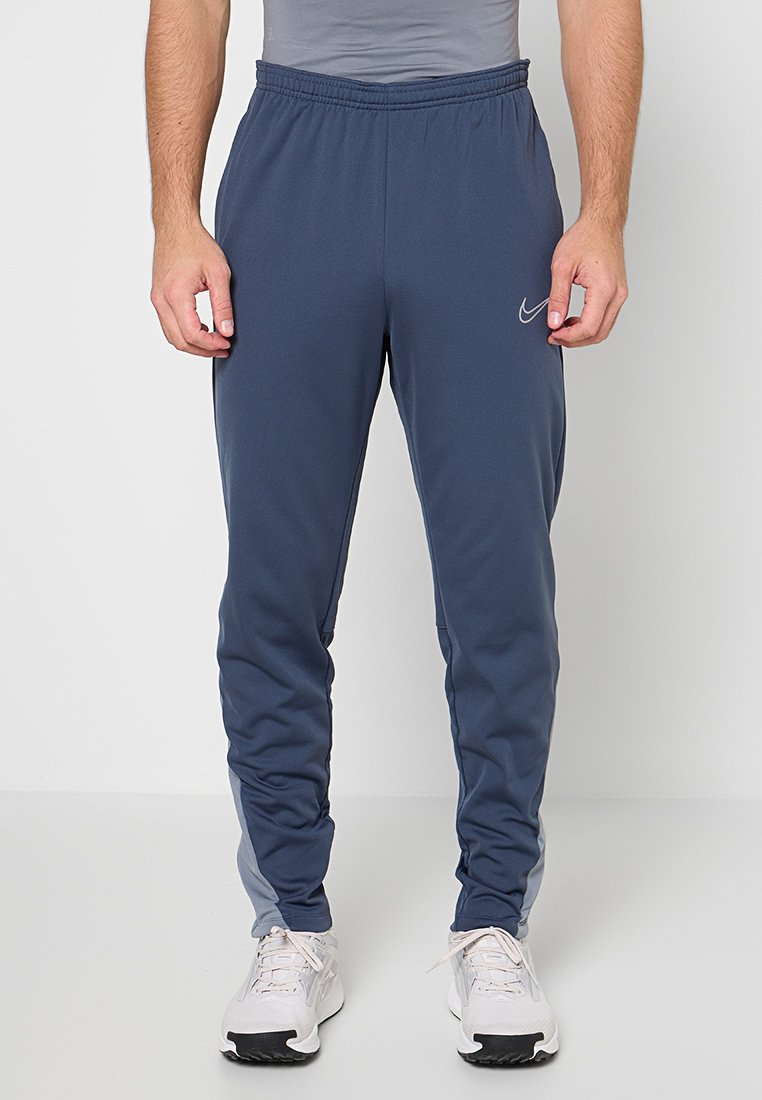 Mann, der blaue Nike-Trainingshose und weiße Laufschuhe trägt, steht vor einem schlichten hellen Hintergrund.