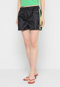 Frau in schwarzen gemusterten Shorts mit neongrünen Seitenstreifen, einem grünen cropped Top und roten Absatzsandalen, stehend vor einem weißen Hintergrund.