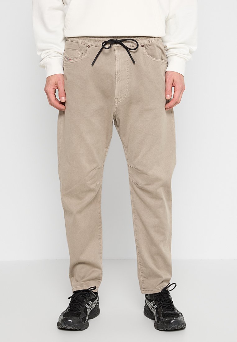 Beige Freizeit-Hosen mit Fronttaschen und schwarzem Kordelzug, getragen von einer Person in schwarzen Sneakers und einem weißen Sweatshirt.
