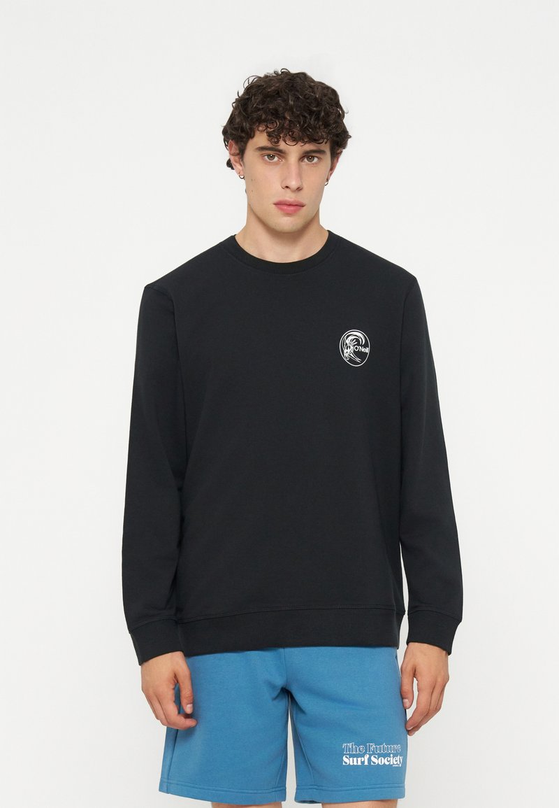 Junger Mann mit lockigem Haar, der ein schwarzes Langarmshirt und blaue Shorts mit dem Schriftzug ‚The Future Surf Society‘ am linken Bein trägt.