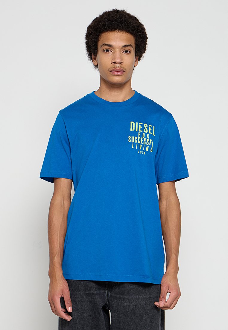 Blaues Baumwoll-T-Shirt mit kurzen Ärmeln, rundem Halsausschnitt und einem grafischen Aufdruck in gelbem Text auf der linken Seite. Kombiniert mit dunkler Hose.