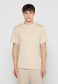 Junger Mann steht aufrecht, trägt ein schlichtes beiges T-Shirt und passende beige Hosen vor einem einfarbigen hellen Hintergrund.
