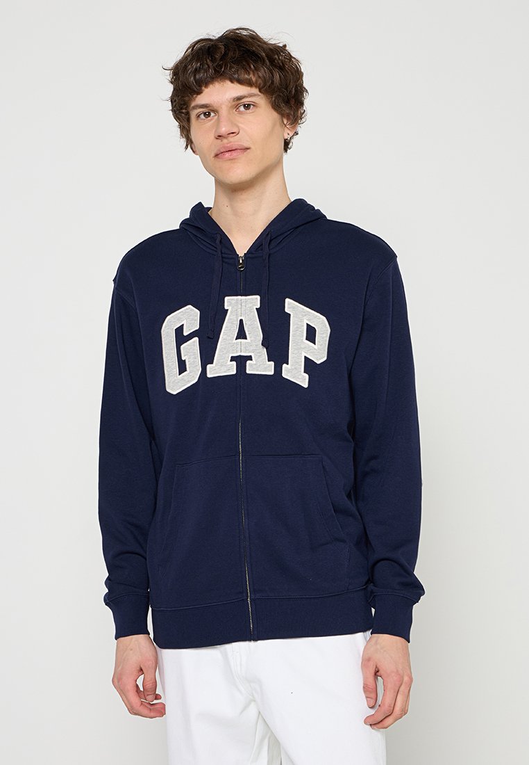 Junger Mann trägt einen marineblauen Kapuzenpullover mit Reißverschluss und dem Schriftzug "GAP" sowie weiße Hose, steht vor einem schlichten hellen Hintergrund.