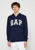Junger Mann trägt einen marineblauen Kapuzenpullover mit Reißverschluss und dem Schriftzug "GAP" sowie weiße Hose, steht vor einem schlichten hellen Hintergrund.