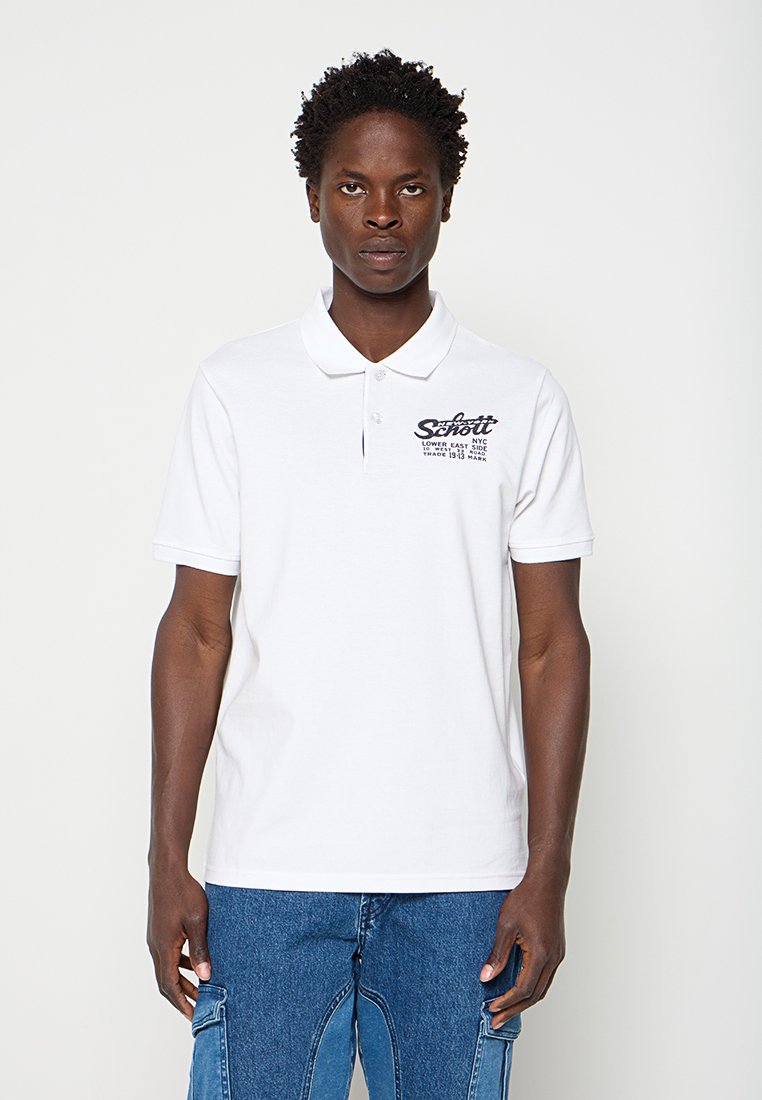 Mann trägt ein weißes Poloshirt mit Text auf der linken Brustseite und blaue Denim-Cargohosen, steht vor einem einfarbigen hellen Hintergrund.