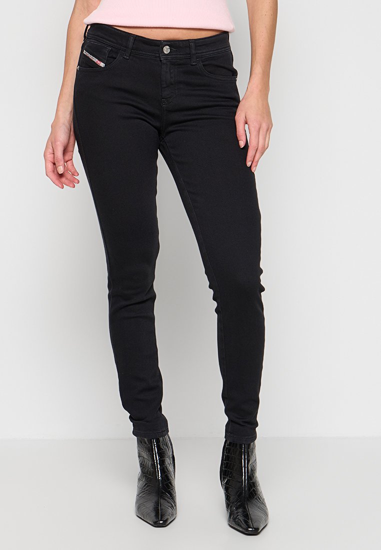 Frau trägt schwarze Skinny-Jeans und schwarze spitze Stiefeletten, steht vor einem weißen Hintergrund.