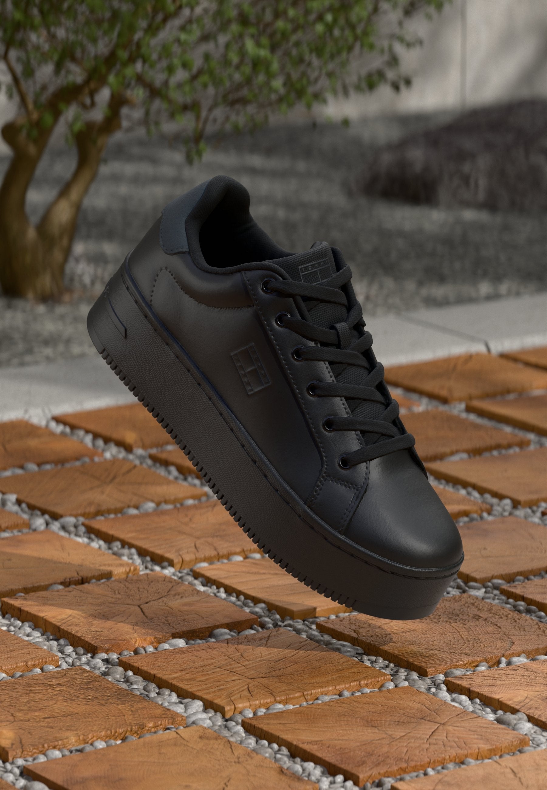Schwarze Sneaker aus glattem Leder mit runder Zehenkappe, Schnürdesign, flacher Sohle und strukturierten Akzenten. Der Hintergrund zeigt braune Fliesen und Kieselsteine.