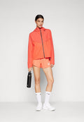 Helle korallenfarbene Sportjacke mit Kapuze und durchgehendem Reißverschluss, kombiniert mit passenden Shorts und einer schwarzen Wasserflasche. Weiße Sneakers und Socken runden das Outfit ab.