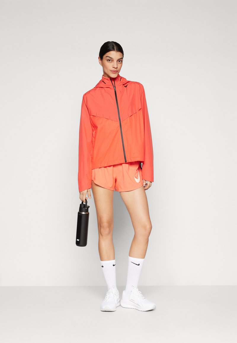 Helle korallenfarbene Sportjacke mit Kapuze und durchgehendem Reißverschluss, kombiniert mit passenden Shorts und einer schwarzen Wasserflasche. Weiße Sneakers und Socken runden das Outfit ab.