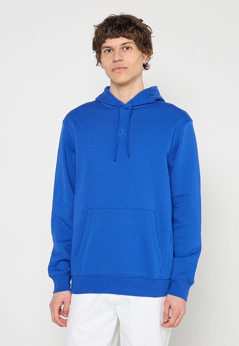 Junger Mann trägt einen blauen Kapuzenpullover mit Fronttasche und weiße Hose, steht vor einem schlichten hellen Hintergrund.