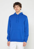 Junger Mann trägt einen blauen Kapuzenpullover mit Fronttasche und weiße Hose, steht vor einem schlichten hellen Hintergrund.