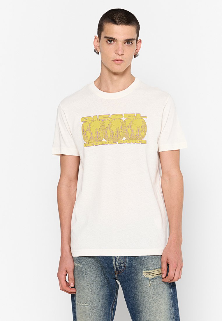 Junger Mann, der ein weißes Grafik-T-Shirt mit gelbem "Diesel Global Dance"-Text und blauen verwaschenen Jeans trägt, steht vor einem weißen Hintergrund.