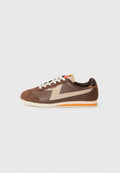 Braune Sneakers aus Leder und Wildleder, mit einem beigen dekorativen Streifen und einer strukturierten braunen Zehenkappe. Weiße Schnürsenkel und orangefarbene Akzente.