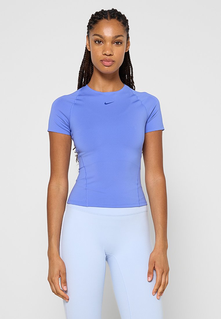 Junge Frau mit langem geflochtenem Haar trägt ein enges hellblaues kurzärmeliges Sportshirt und blassblaue Leggings und steht vor einem einfarbigen Hintergrund.