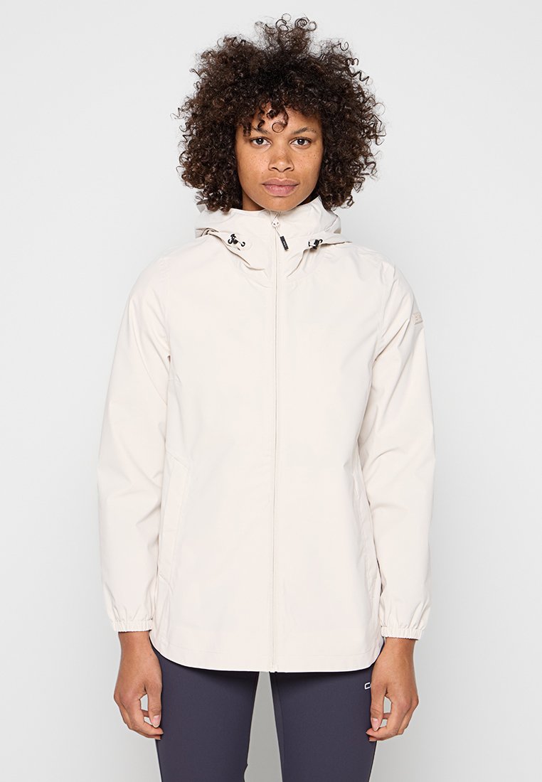 Frau mit lockigem Haar, die eine hellbeige Kapuzenjacke und dunkle Leggings trägt, steht vor einem schlichten hellen Hintergrund.