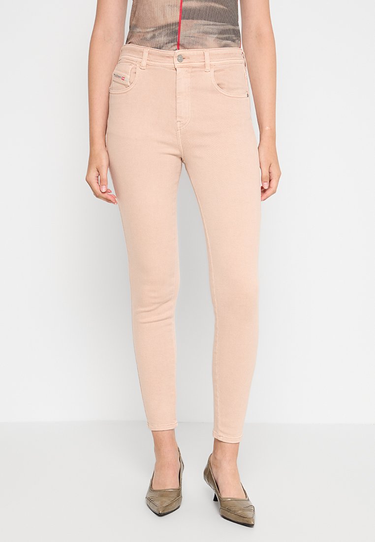 Eine Frau trägt hellrosa enge Jeans und metallische spitze Absatzschuhe, während sie vor einem schlichten weißen Hintergrund steht.